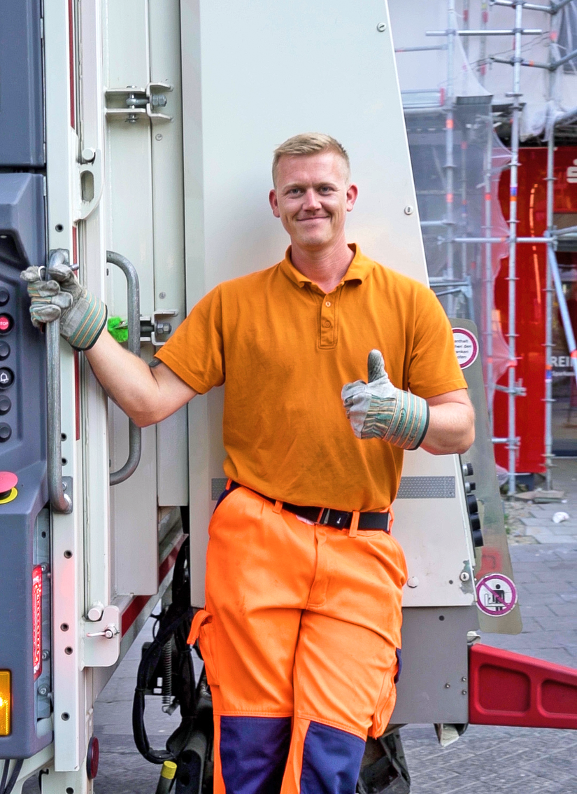 Junger Mann steht auf dem Brett hinter dem Entsorgungsfahrzeug und zeigt mit dem Daumen nach oben