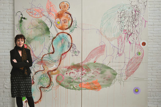 Seet van Hout vor einem ihrer Großformaten, welches an eine weiße Wand gehangen ist. Sie schaut in die Kamera und trägt schwarze Kleidung.