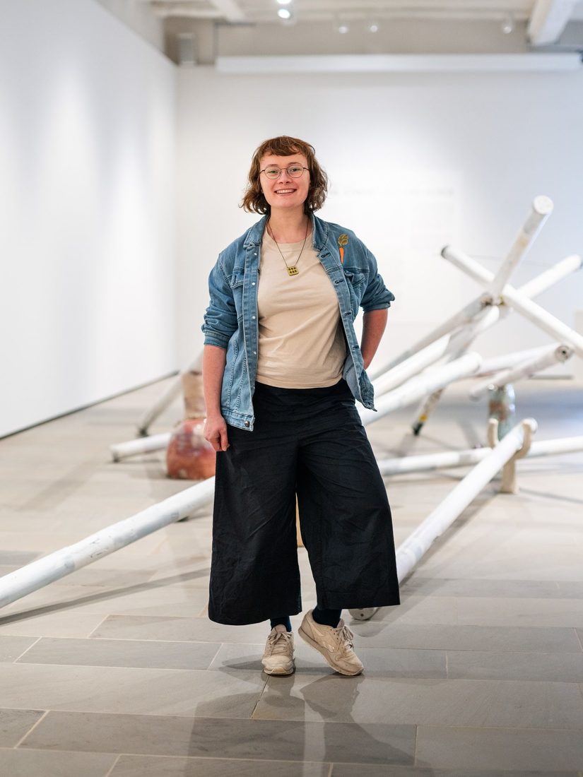 Irene Strese, eine junge weiße Frau, steht vor einem Kunstwerk aus Reithürden in der Galerie und schaut in die Kamera.