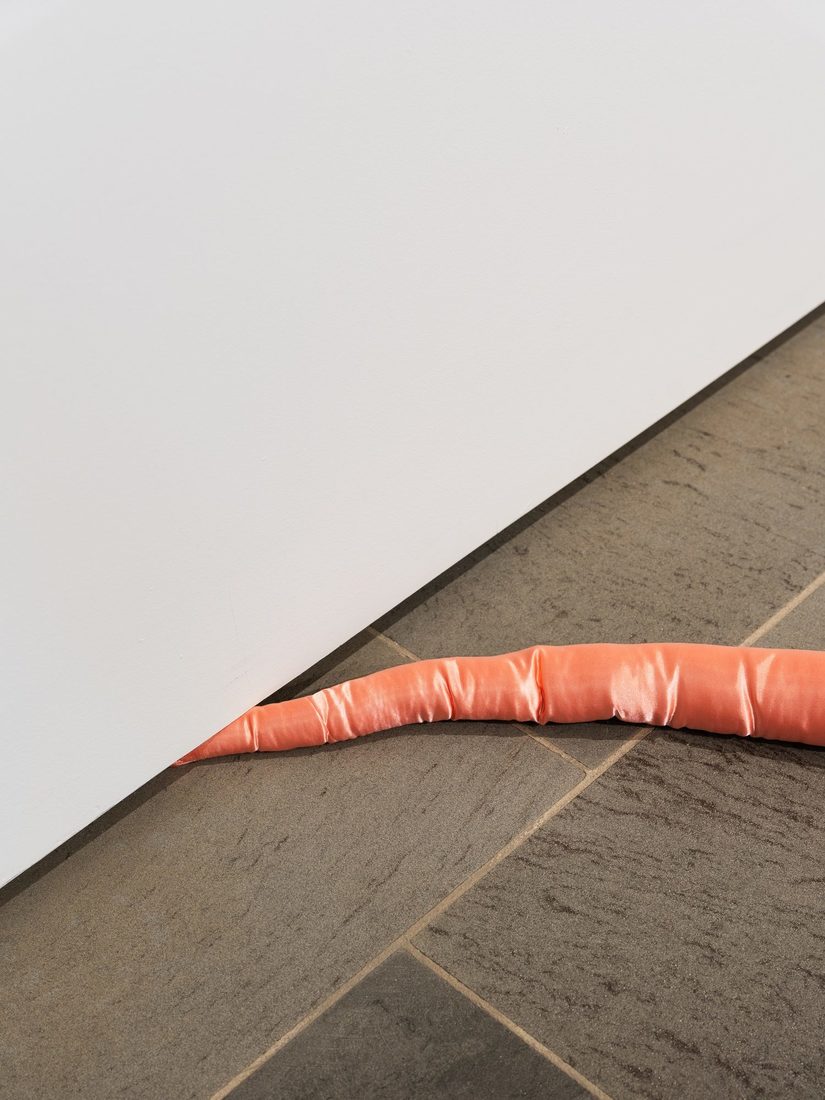 Grauer Fliesenboden mit weißer Stellwand. Eine rosane, runde Stoffrolle verschwindet unte der Stellwand.