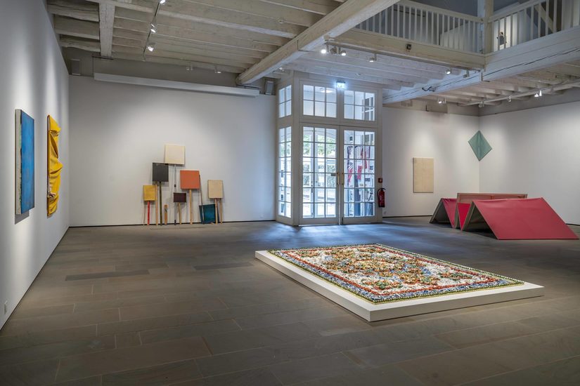 Blick auf den Eingangsbereich der Reithalle, an der Wand links und vor Kopf hängen verschiedenfarbige Leinwände. Rechts hinten in der Ecke steht ein Konstrukt aus pinken Leinwänden, mittig im Raum liegt ein florarer Teppich aus einzelnen Scherben.