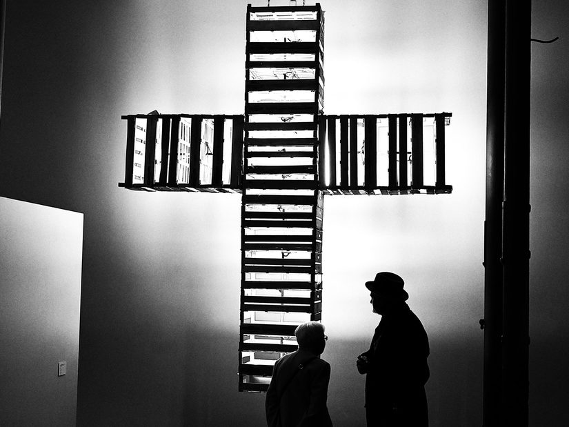 Installation: Einfache Obstkisten sind zu einem gigantischen Holzkreuz zusammen gebaut worden. Sie sind ausgestattet mit Neonröhren. Das Kreuz hängt von der Decke und leuchtet in Streifen aus den Lücken der Obstkisten heraus.