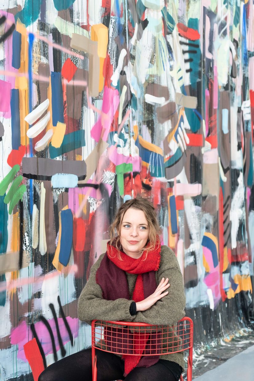 Nina Hanna Kornatz sitzt auf einem roten Metallstuhl. Sie blickt rechts an der Kamera vorbei. Im Hintergrund ist eine bunte bemalte Stoffbahn.