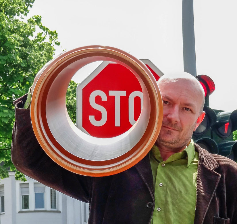 Ruppe Koselleck schaut frontal in die Kamera. Auf der linken Schulter trägt er ein großes orangenes Kunststoffrohr. Im Hintergrund sind eine Ampel und ein Stopschild.