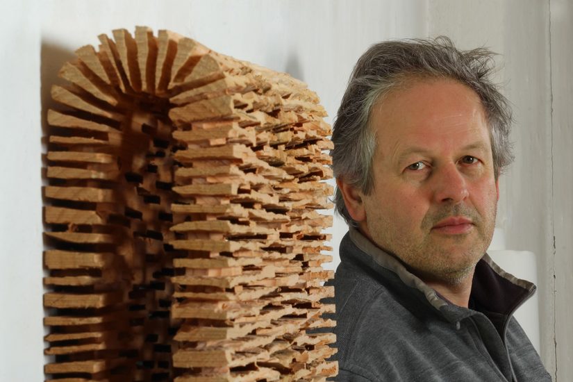 Stephan Wurmer steht rechts neben einer Holzskulptur und blickt frontal in die Kamera.