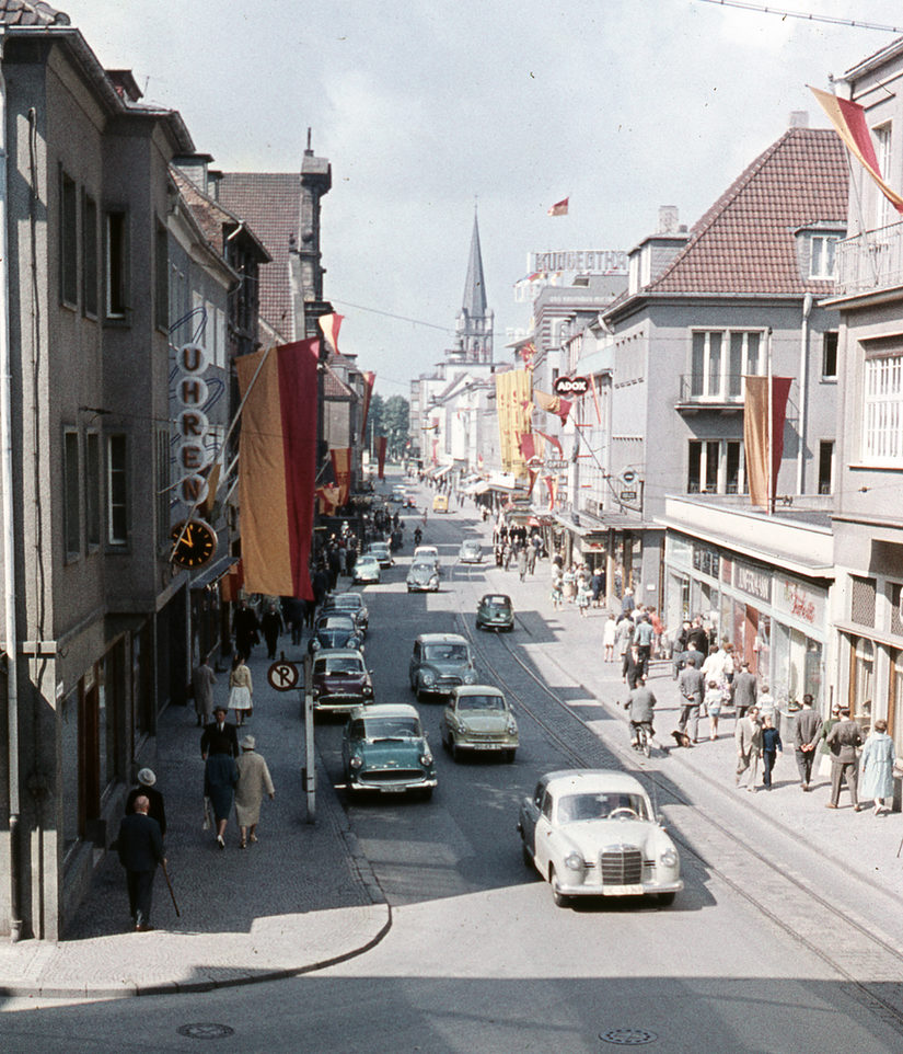 Straße mit vielen Autos und Menschen. Im Hintergrund der Dom.