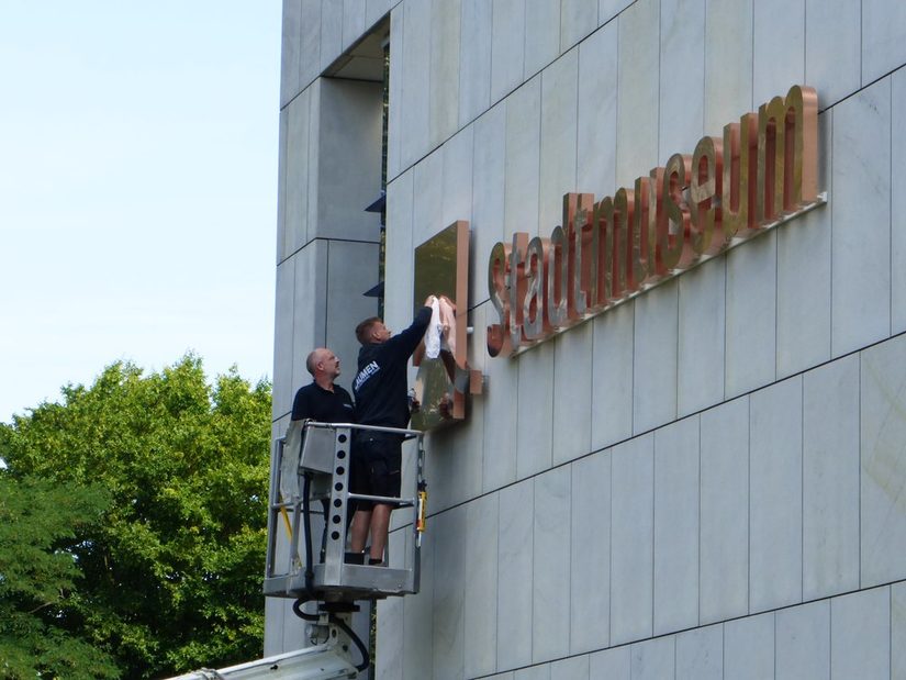 Fassade mit der Aufschrift Stadtmuseum und zwei weiße Männer, die es auf einen Hubwagen putzen