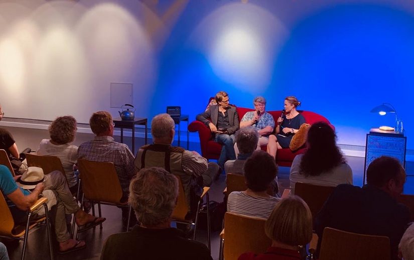 Ein Publikum und drei Menschen die auf einem Sofa sitzen und sie haben Mikrofone in der Hand.