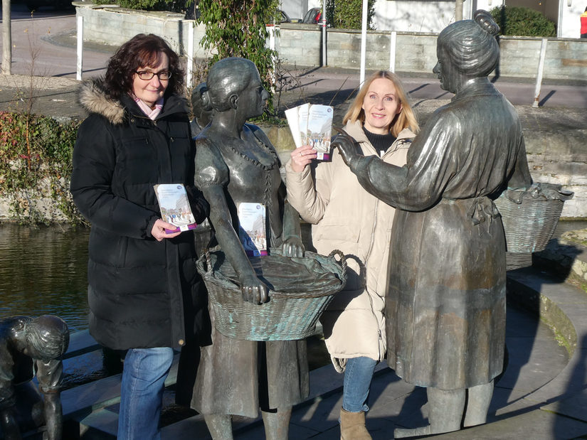 Ingrid Vogedes (li.) und Alexandra Bröckling vom „Team Gästeführungen“ der Tourist Information Paderborn präsentieren die neue Broschüre „Öffentliche Führungen 2024“ beim Waschfrauendenkmal im Paderquellgebiet, das bei fast jeder Stadtführung gezeigt wird.